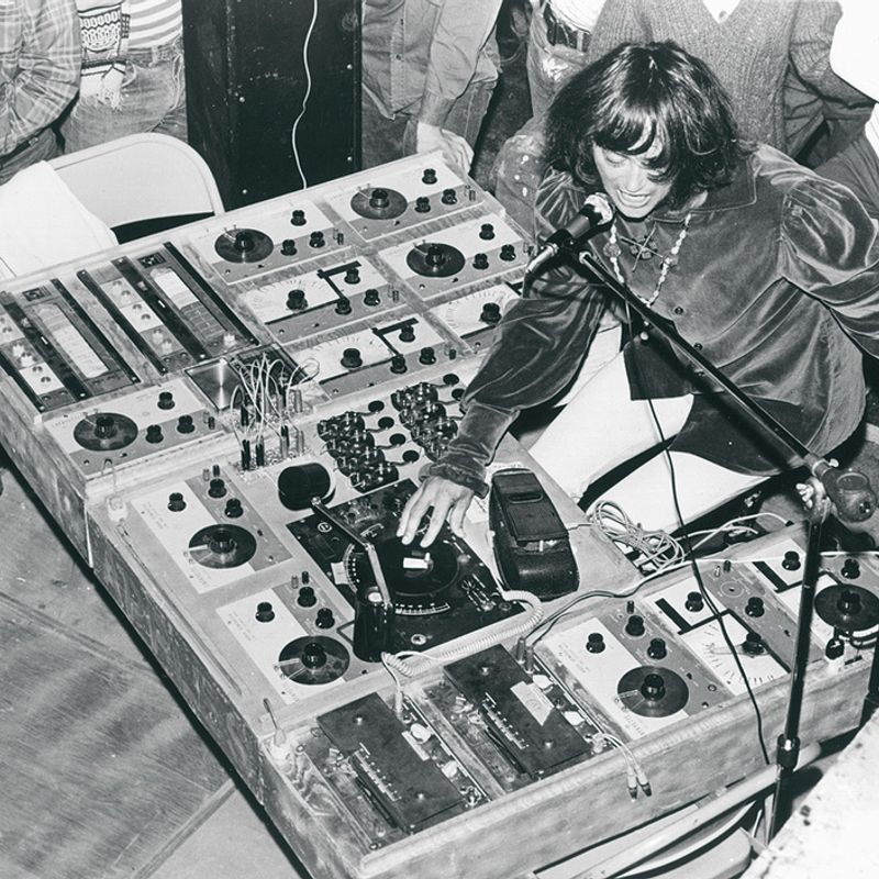 A black and white film photograph of a young Simeon of Silver Apples singing into a microphone and manipulating a complicated looking selection of audio oscillator machines.