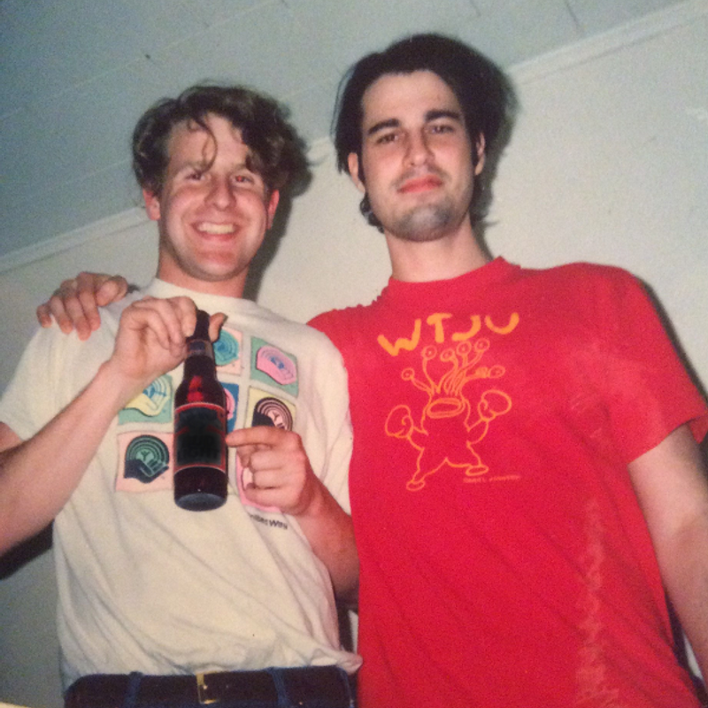 A colour film photograph of a young David Berman with his arm around a young Bob Nastanovich (both of the band Silver Jews). Nastanovich is holding and pointing to a beer bottle. Berman is wearing a red WTJU tee shirt with art that looks like it's one of Daniel Johnston's frog like creatures but with many eyes on stalks.